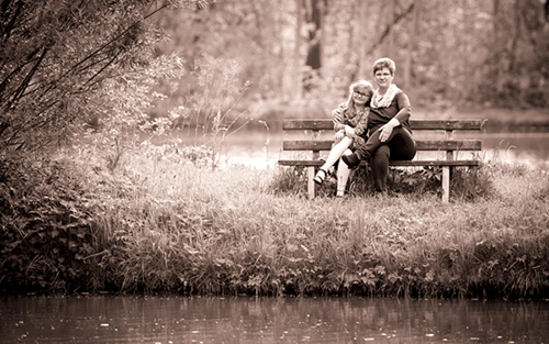 Sepia-farbenes Porträt einer Mutter mit ihrer Tochter, die eng umschlungen auf einer Holzbank am Ufer eines Gewässers in der Natur sitzen. Ein herzliches Mutter-Tochter-Fotoshooting, stimmungsvoll festgehalten von Michael Schalansky Fotografie in Erfurt.